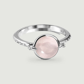 A silver ring with an oval pink stone and small clear accents rests on a light gray surface.