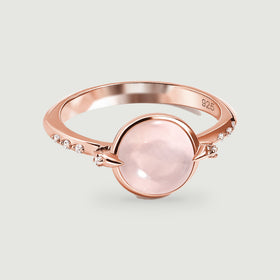 A rose gold ring with a rounded pink stone is centered above a soft shadow on light gray.
