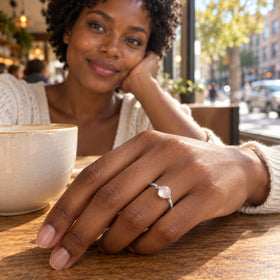 The silver ring is worn on a hand resting beside a ceramic cup on a wooden cafe table.