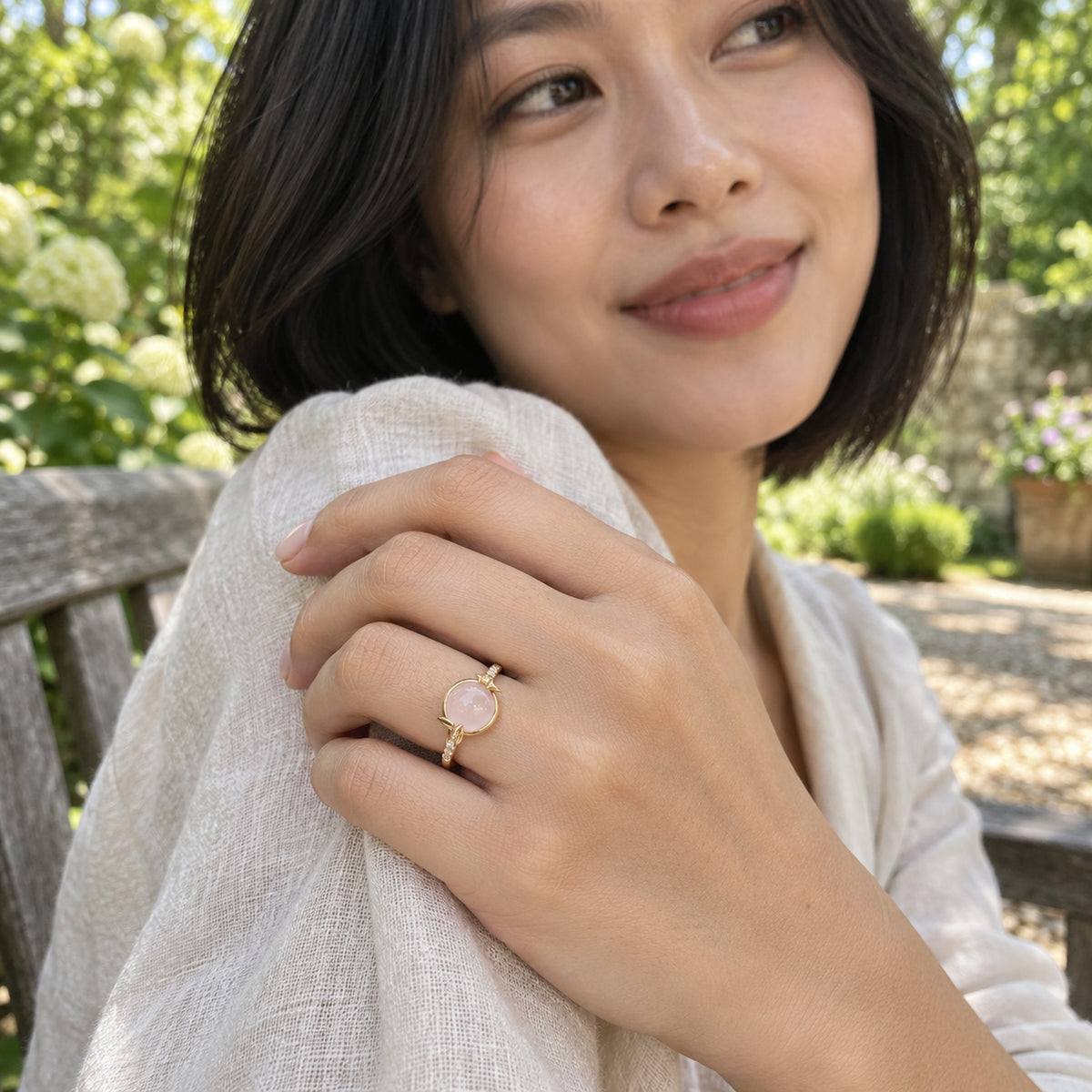 A yellow gold ring with a pink cabochon is worn on a hand holding a linen sleeve beside a garden bench.