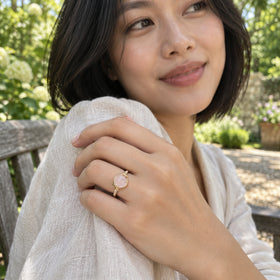 A yellow gold ring with a pink cabochon is worn on a hand holding a linen sleeve beside a garden bench.