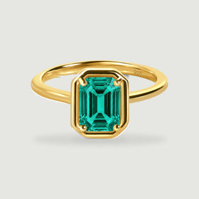 A yellow gold vermeil ring with a rectangular green stone is centered on a light grey surface.