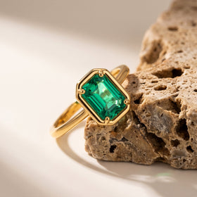 A yellow gold ring with a rectangular green stone rests against textured beige stone.