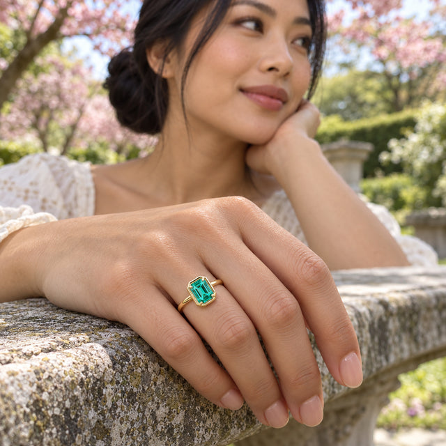 A woman shows the yellow gold green-stone ring on her hand resting on a stone garden bench.