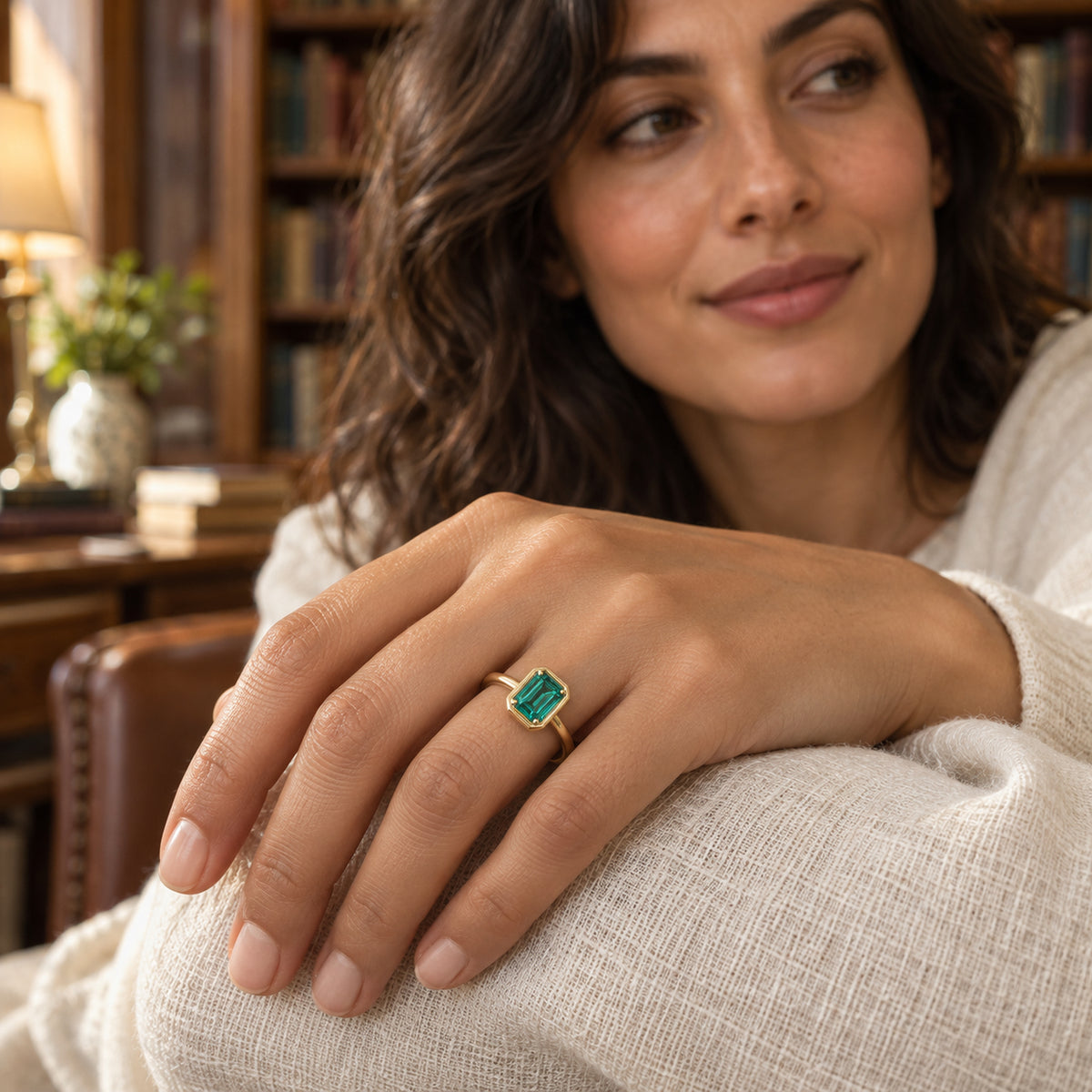 A yellow gold ring with a rectangular green stone is worn on a hand resting on an ivory linen sleeve in a reading room.