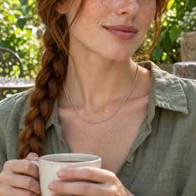 A silver chain is worn with an open sage linen shirt on a leafy patio.