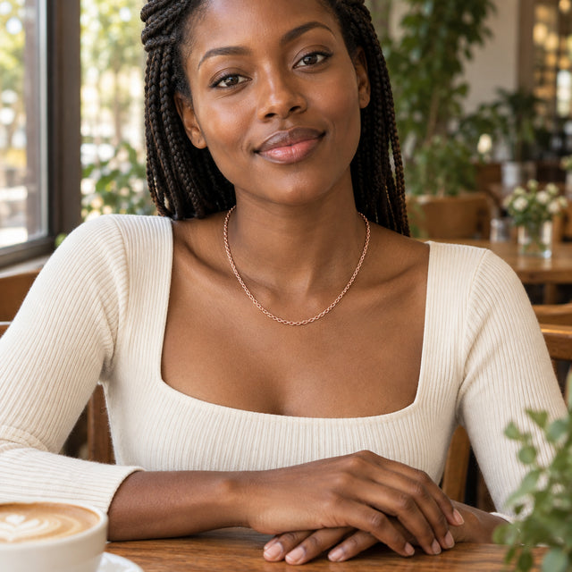A woman in a cream top wears a rose gold chain at a wooden cafe table.