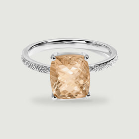 A rectangular champagne quartz is held by four silver prongs above a leaf-engraved band on a pale gray surface.