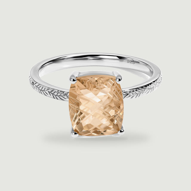 A rectangular champagne quartz is held by four silver prongs above a leaf-engraved band on a pale gray surface.