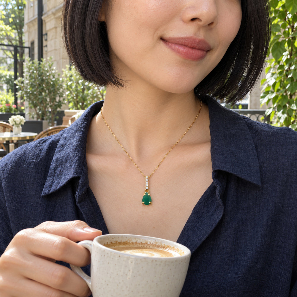 A woman in a navy blouse wears the green pendant on a yellow gold chain while holding a cup at a leafy cafe.