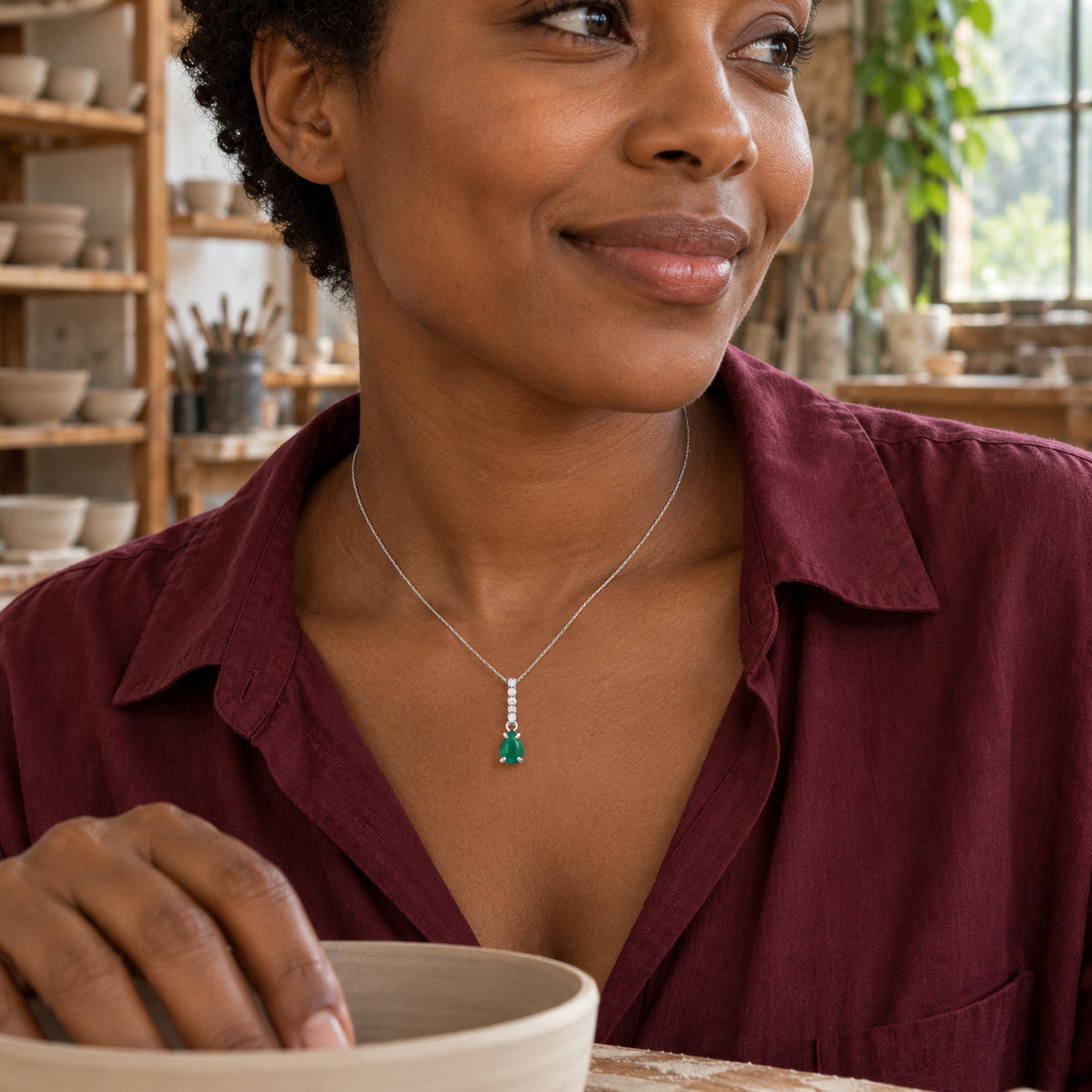 A woman in a burgundy shirt wears the silver green-stone necklace while holding a ceramic bowl in a pottery workshop.