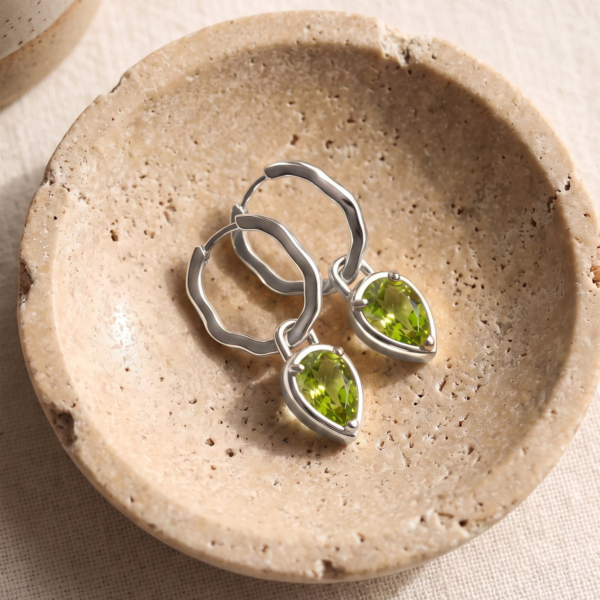 Silver-tone peridot hoops lie together on a round beige stone dish over woven fabric.