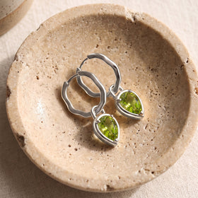 Silver-tone peridot hoops lie together on a round beige stone dish over woven fabric.