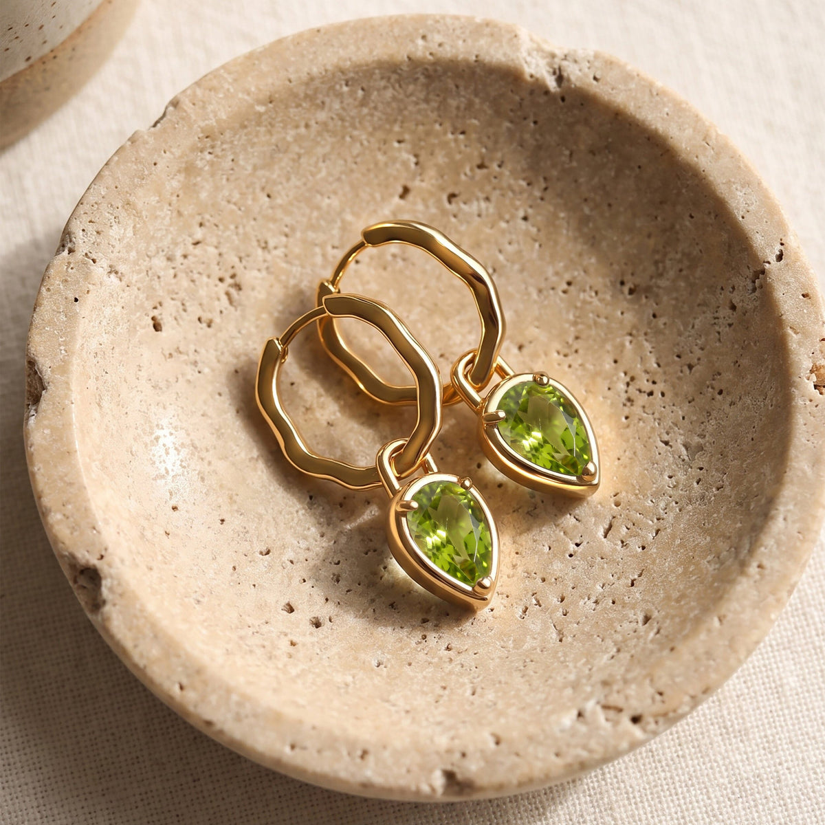 Two yellow gold-tone earrings with green teardrop stones rest on a textured stone tray.