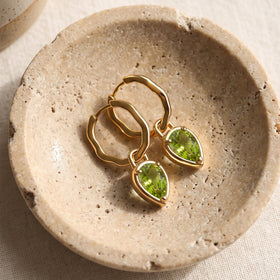 Two yellow gold-tone earrings with green teardrop stones rest on a textured stone tray.