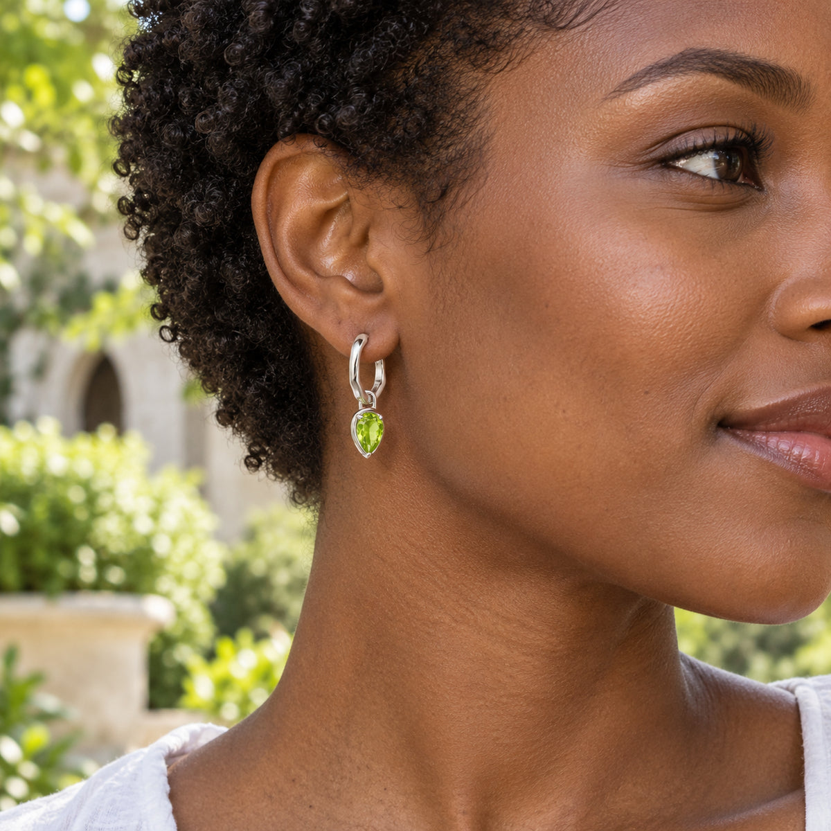 A woman with short dark curls wears a silver-tone peridot hoop earring in a leafy courtyard.