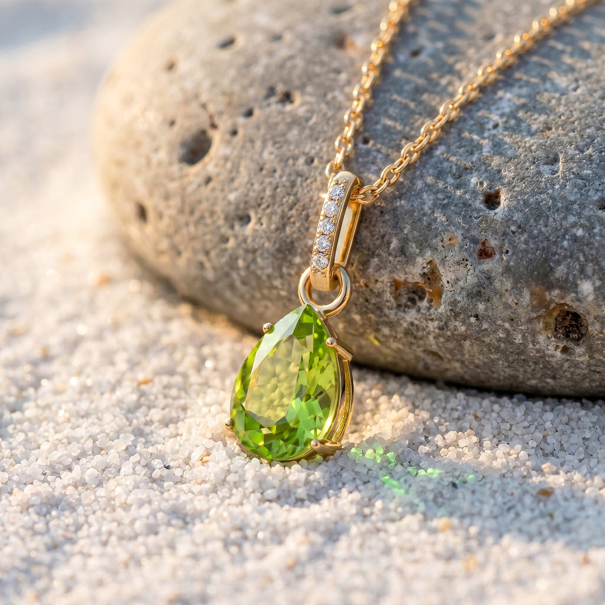 A yellow gold necklace rests against a textured gray pebble on pale sand with its green pear pendant in sunlight.