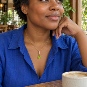 A woman with short coiled hair wears the yellow gold peridot necklace with a cobalt blue shirt beside a cafe window.