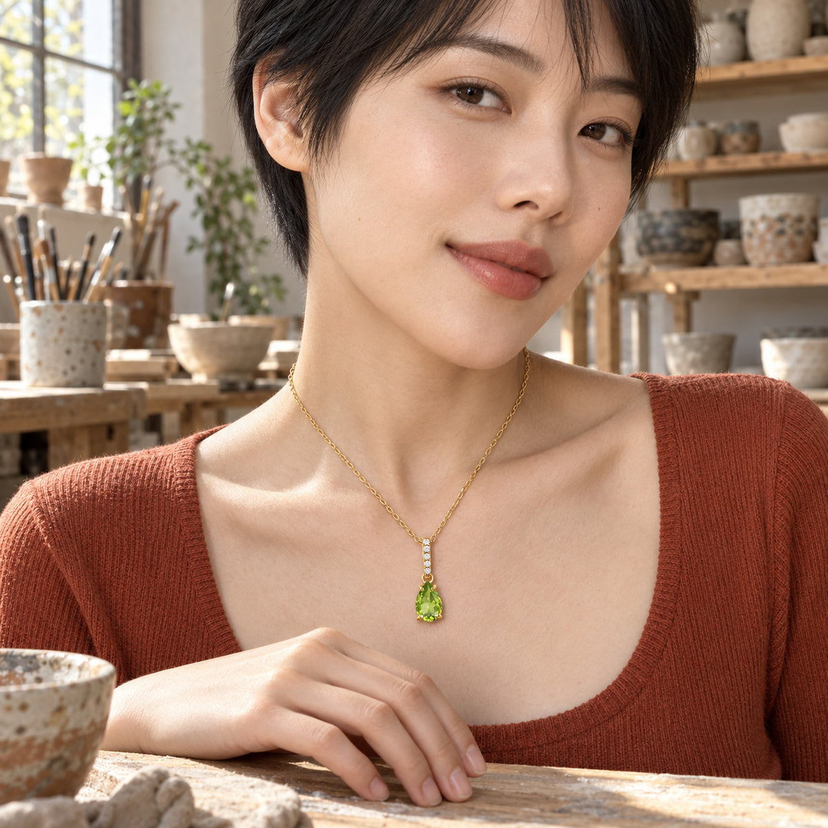 A green pear pendant on a yellow gold chain rests above a terracotta neckline in a sunlit pottery studio.
