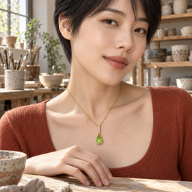 A green pear pendant on a yellow gold chain rests above a terracotta neckline in a sunlit pottery studio.