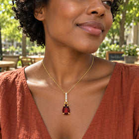 A woman in a rust-colored linen blouse wears the yellow gold garnet necklace at a leafy outdoor cafe.