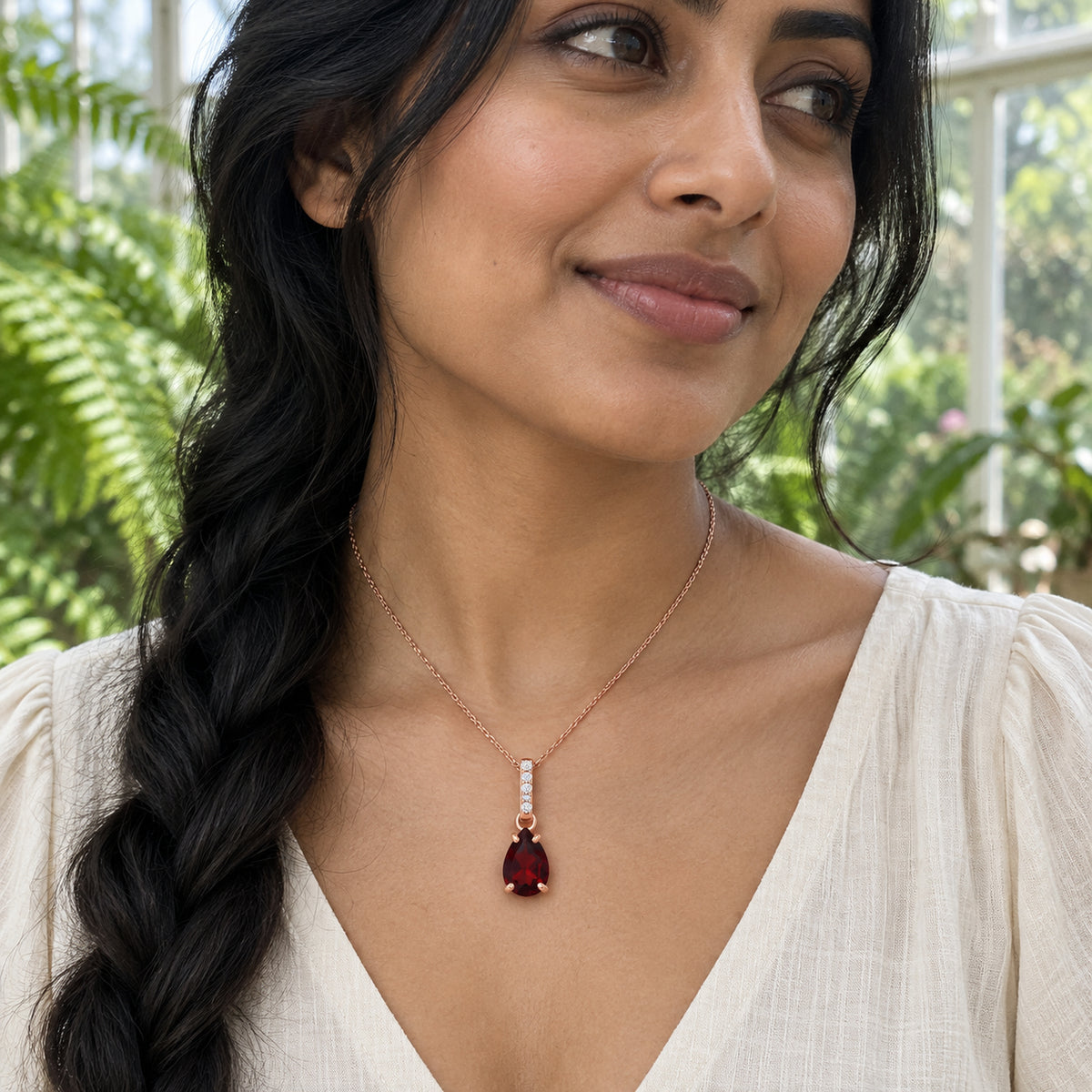 A woman with a long dark braid wears the rose gold garnet pendant with a cream blouse among conservatory plants.