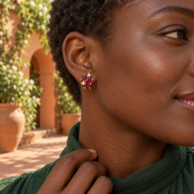 A woman with short natural curls wears a rose gold stud with pink and deep red stones in a sunlit terracotta courtyard.