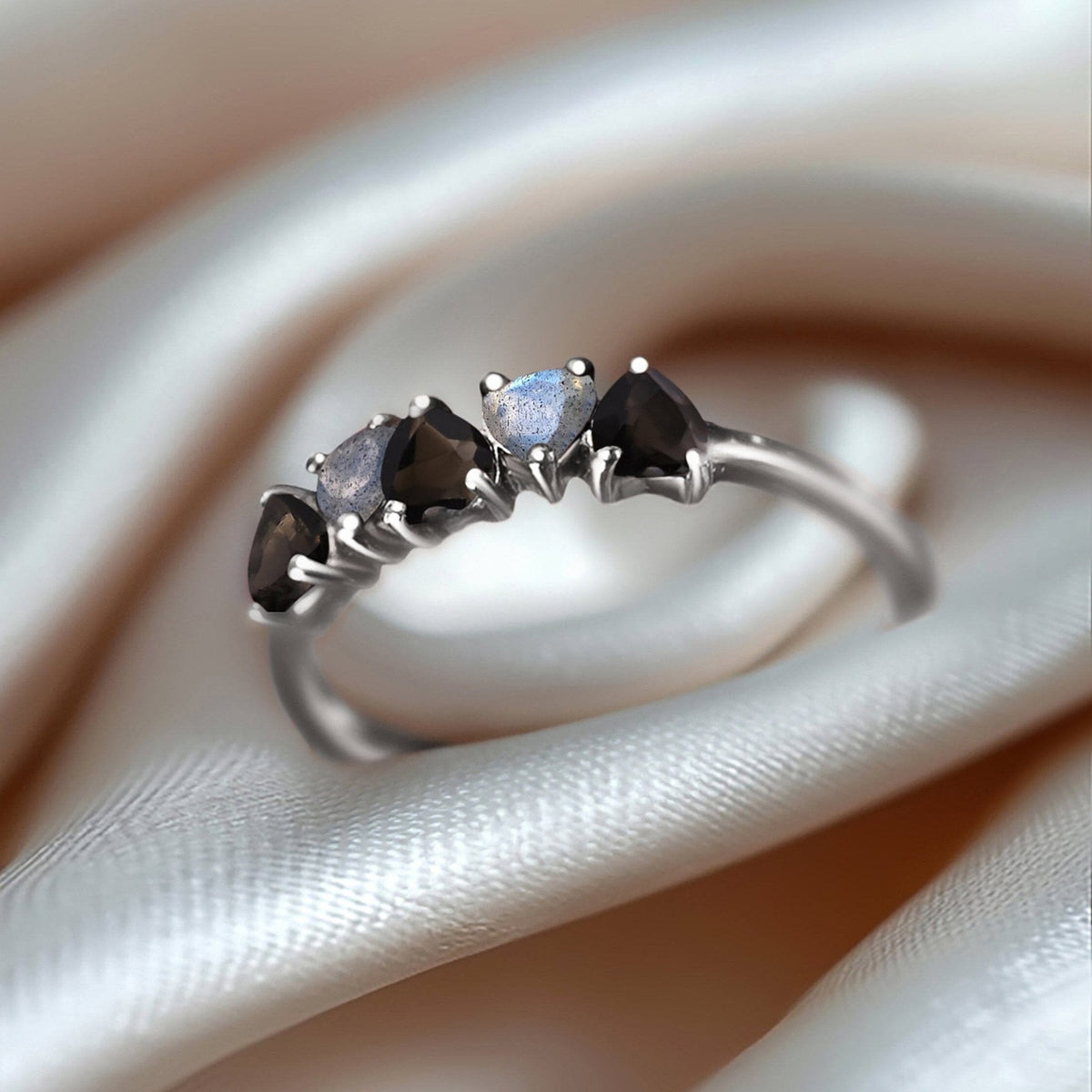 The silver ring rests on folded pale satin with alternating dark and iridescent heart-shaped stones facing the light.