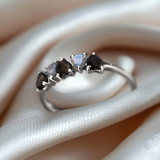 The silver ring rests on folded pale satin with alternating dark and iridescent heart-shaped stones facing the light.