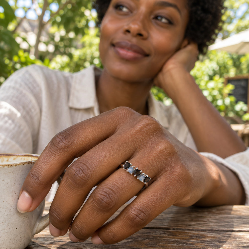 A woman wears the silver ring with alternating black and blue-gray heart-shaped stones beside a ceramic cup at an outdoor cafe.