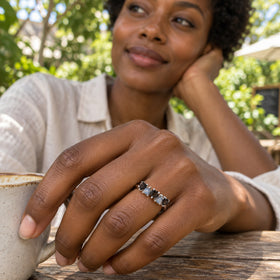 A woman wears the silver ring with alternating black and blue-gray heart-shaped stones beside a ceramic cup at an outdoor cafe.