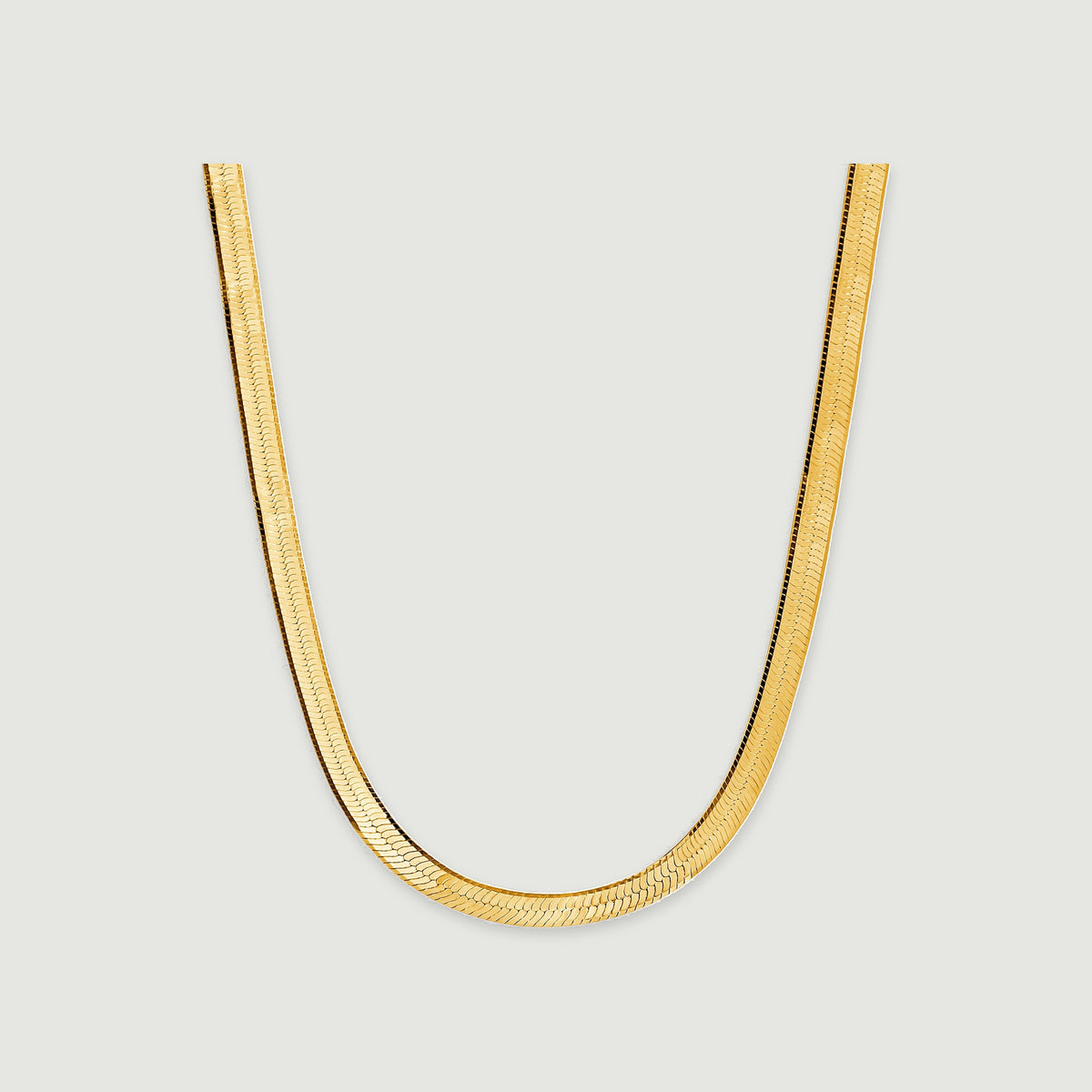 A flat yellow gold herringbone necklace curves into a U shape on a light gray surface.