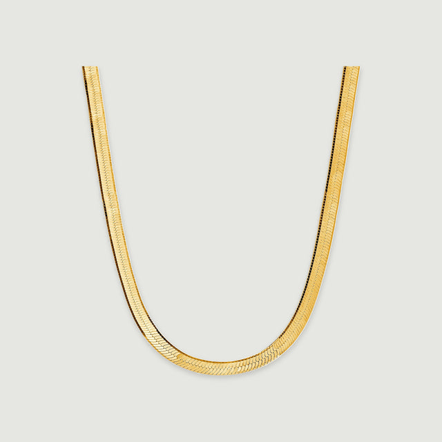 A flat yellow gold herringbone necklace curves into a U shape on a light gray surface.