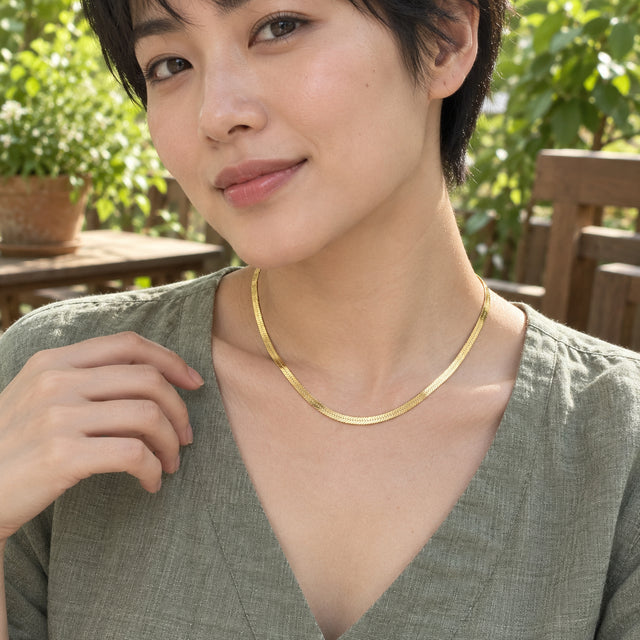 A woman in a sage linen shirt wears the yellow gold herringbone necklace on a leafy garden terrace.
