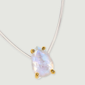 A gold-set moonstone pendant rests at an angle beneath a curved transparent cord on pale gray.