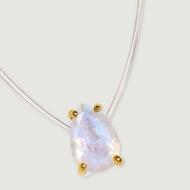 A gold-set moonstone pendant rests at an angle beneath a curved transparent cord on pale gray.