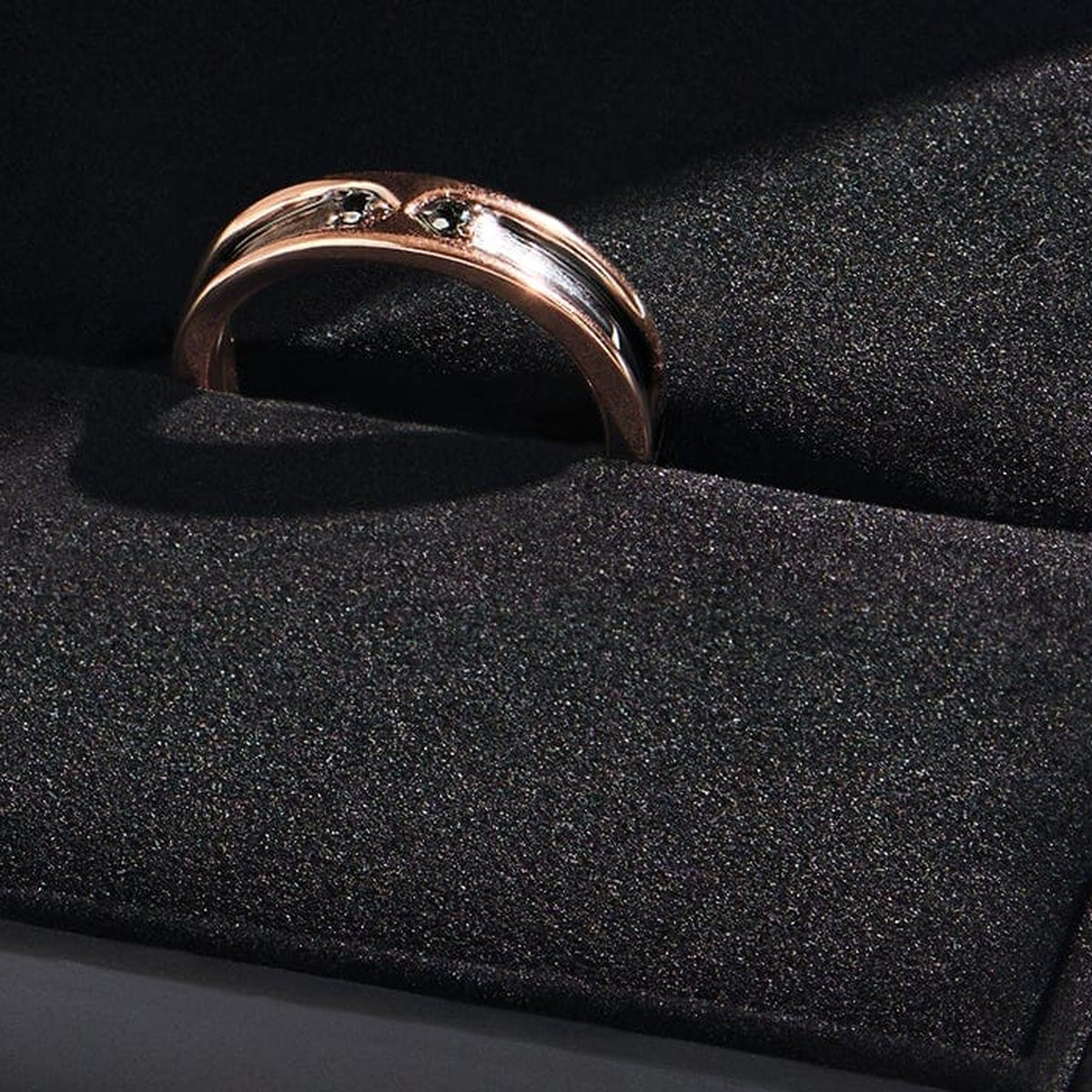 A rose gold ring with two dark stones sits in a slot on a black textured display cushion.