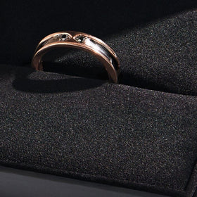 A rose gold ring with two dark stones sits in a slot on a black textured display cushion.