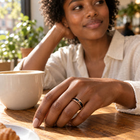 A rose gold ring with a black inlay is worn on a hand beside a ceramic coffee cup on a wooden cafe table.