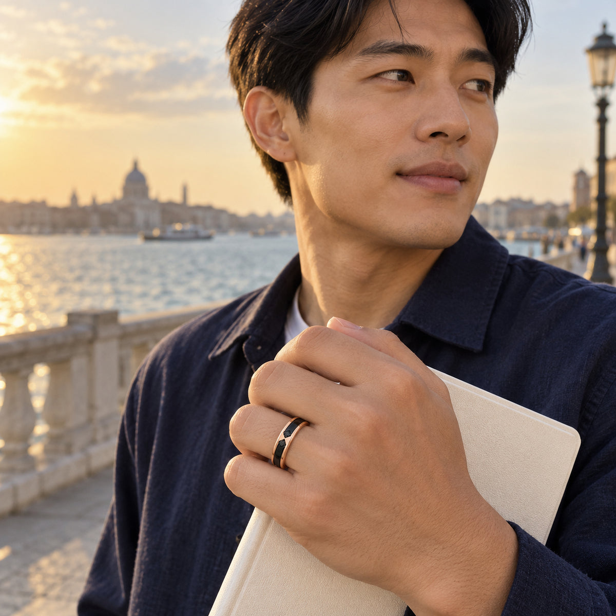 A man wears a rose gold ring with a black inlay while holding a cream notebook beside a waterfront railing.
