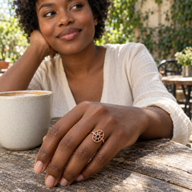 A woman with dark skin wears the rose gold oval cage ring beside a ceramic cup at an outdoor cafe.