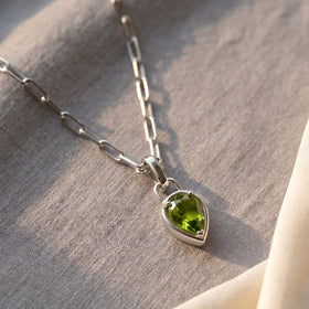 A silver-tone peridot necklace rests diagonally on light gray linen with soft natural shadows.