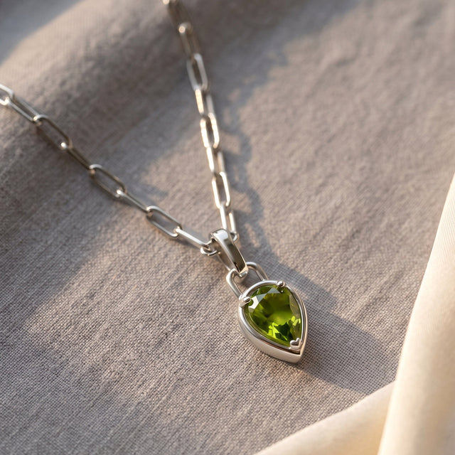 A silver-tone peridot necklace rests diagonally on light gray linen with soft natural shadows.