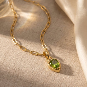 A green pear-shaped pendant and yellow-gold-tone links lie on a pale textured fabric surface.