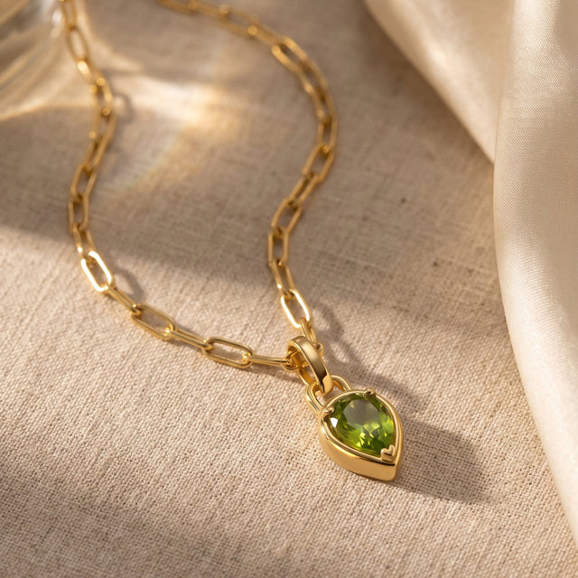 A green pear-shaped pendant and yellow-gold-tone links lie on a pale textured fabric surface.