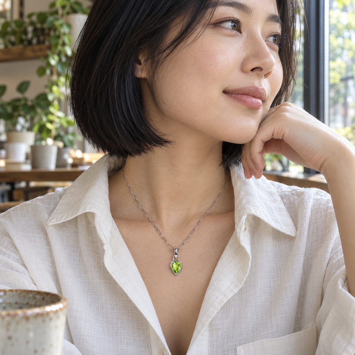 A woman in a white linen shirt wears a silver-tone green peridot pendant beside a cafe window.