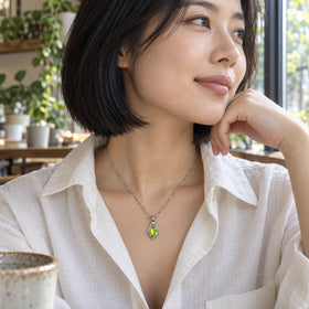 A woman in a white linen shirt wears a silver-tone green peridot pendant beside a cafe window.