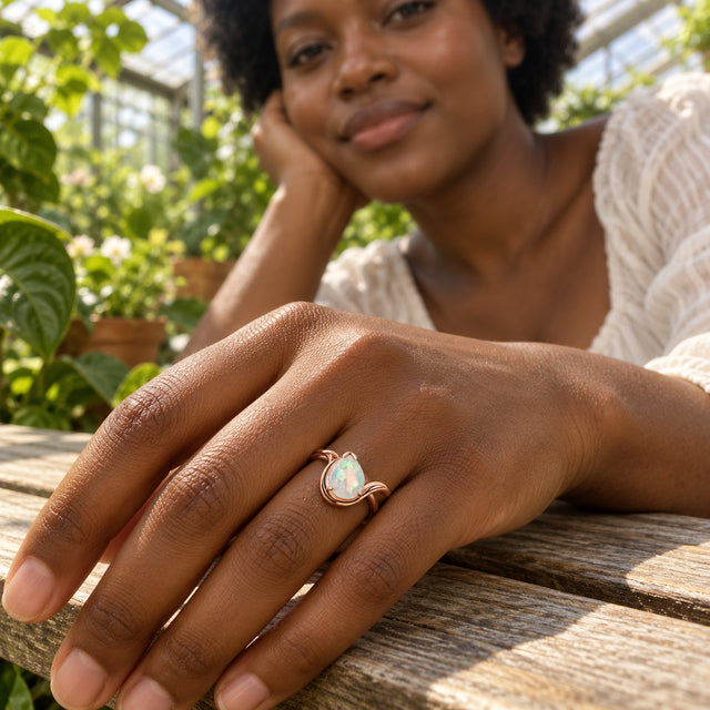 The pear-shaped opal ring is worn on a hand resting on a wooden bench among sunlit greenhouse plants.