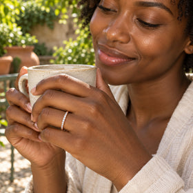 A woman in a cream linen shirt wears a rose gold diamond ring while holding a ceramic cup in a leafy courtyard.