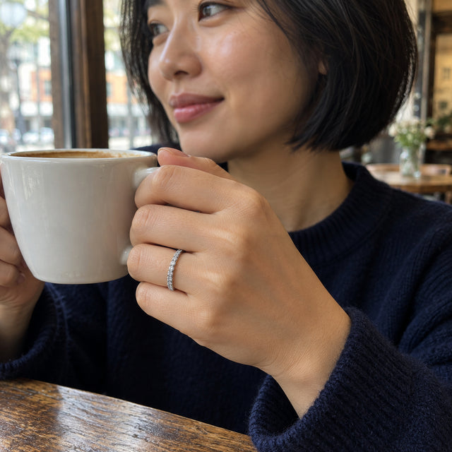 A white gold diamond ring is visible on a hand holding a coffee cup beside a cafe window.